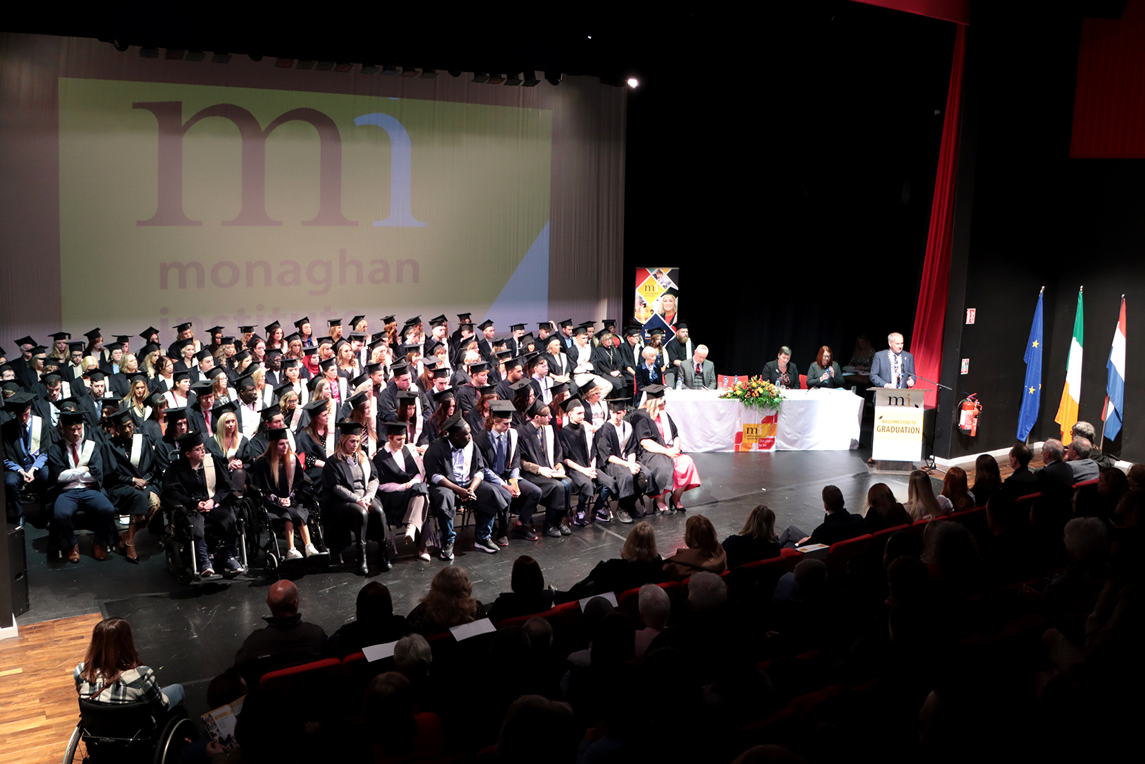 Graduation students sitting with cap and gowns and man speaking on microphone